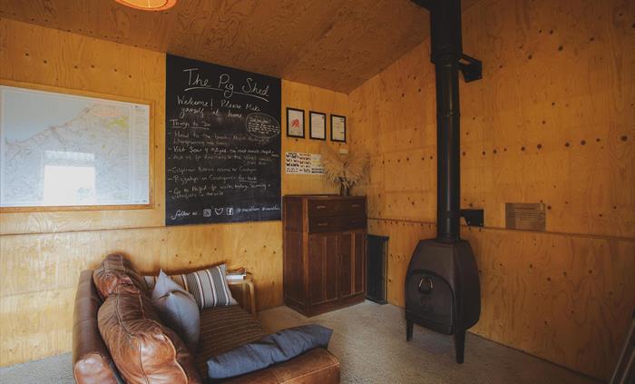 A plywood lined room with a large brown sofa facing a wood burning stove.